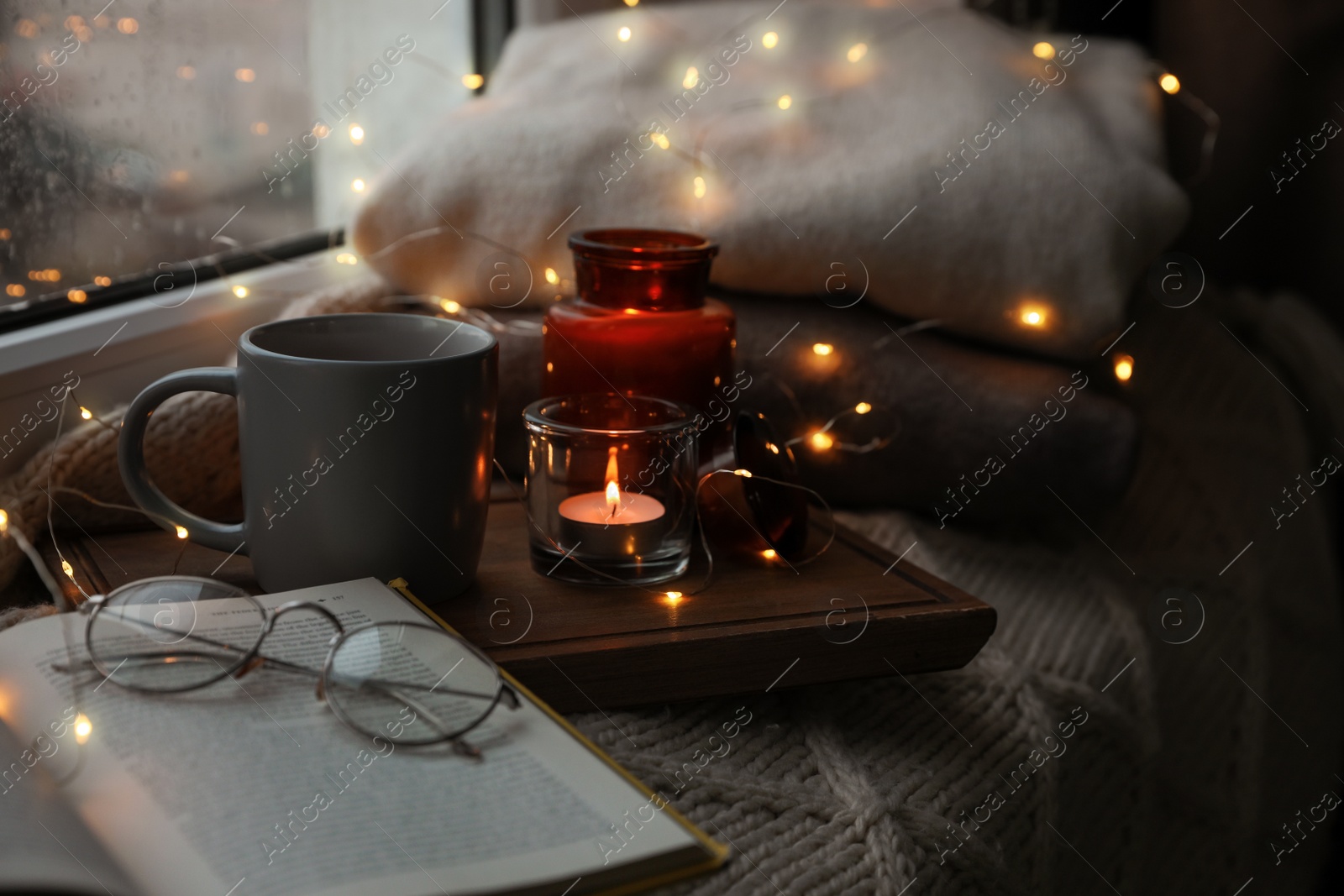 Photo of Cup of hot drink, open book and Christmas lights on knitted blanket near window. Cozy season