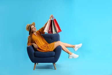 Photo of Beautiful young woman with paper shopping bags in armchair on light blue background