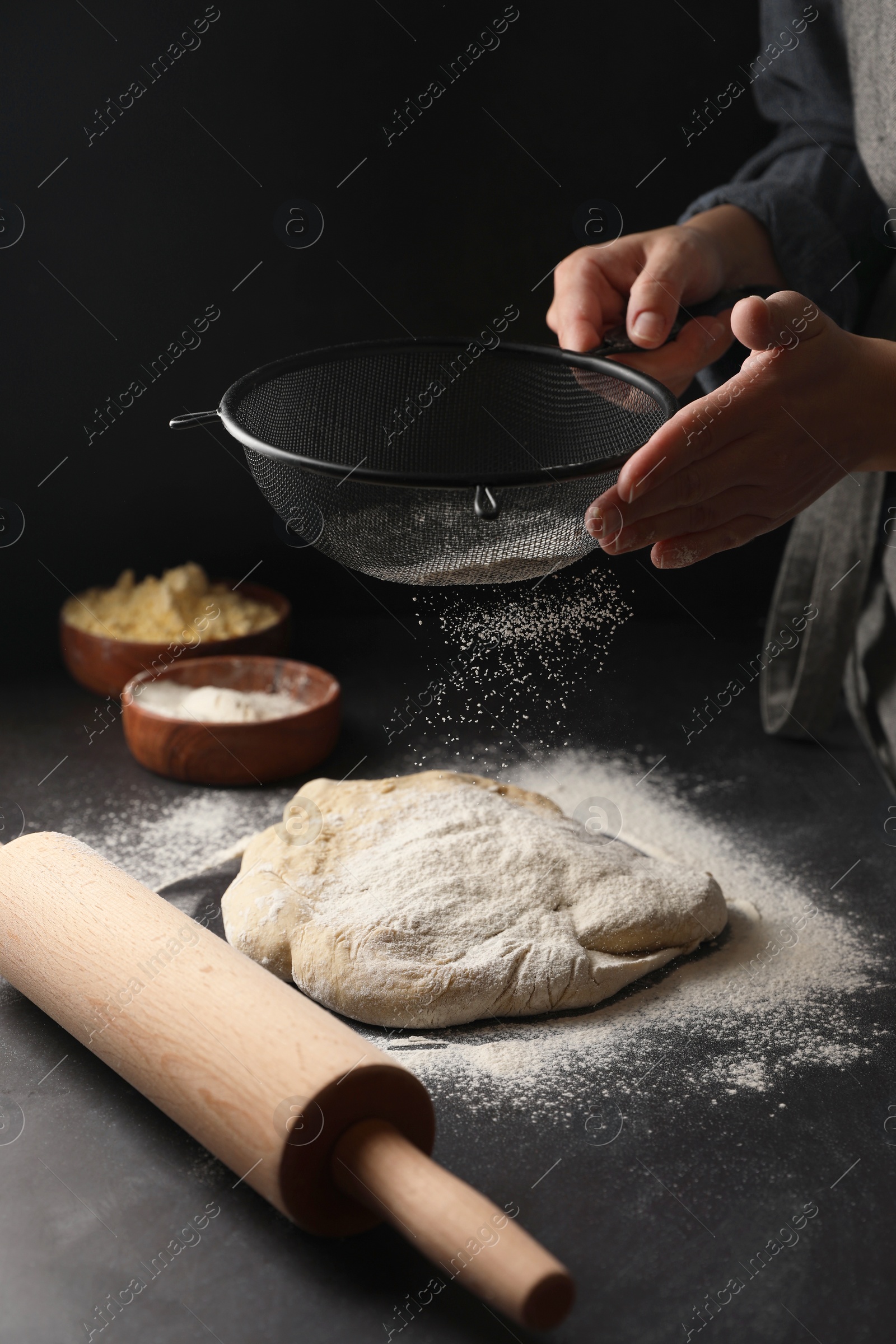 Photo of Woman sprinkling flour over dough at black table, closeup