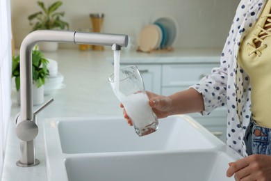 Photo of Woman filling glass with water from tap in kitchen, closeup
