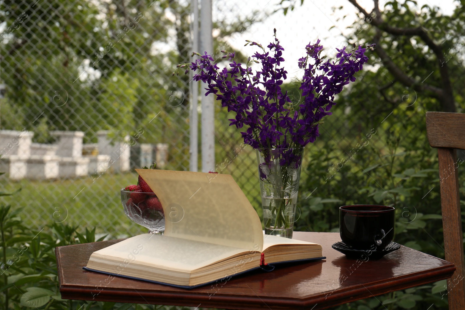 Photo of Open book, cup of tea, strawberries and beautiful wildflowers on table in garden