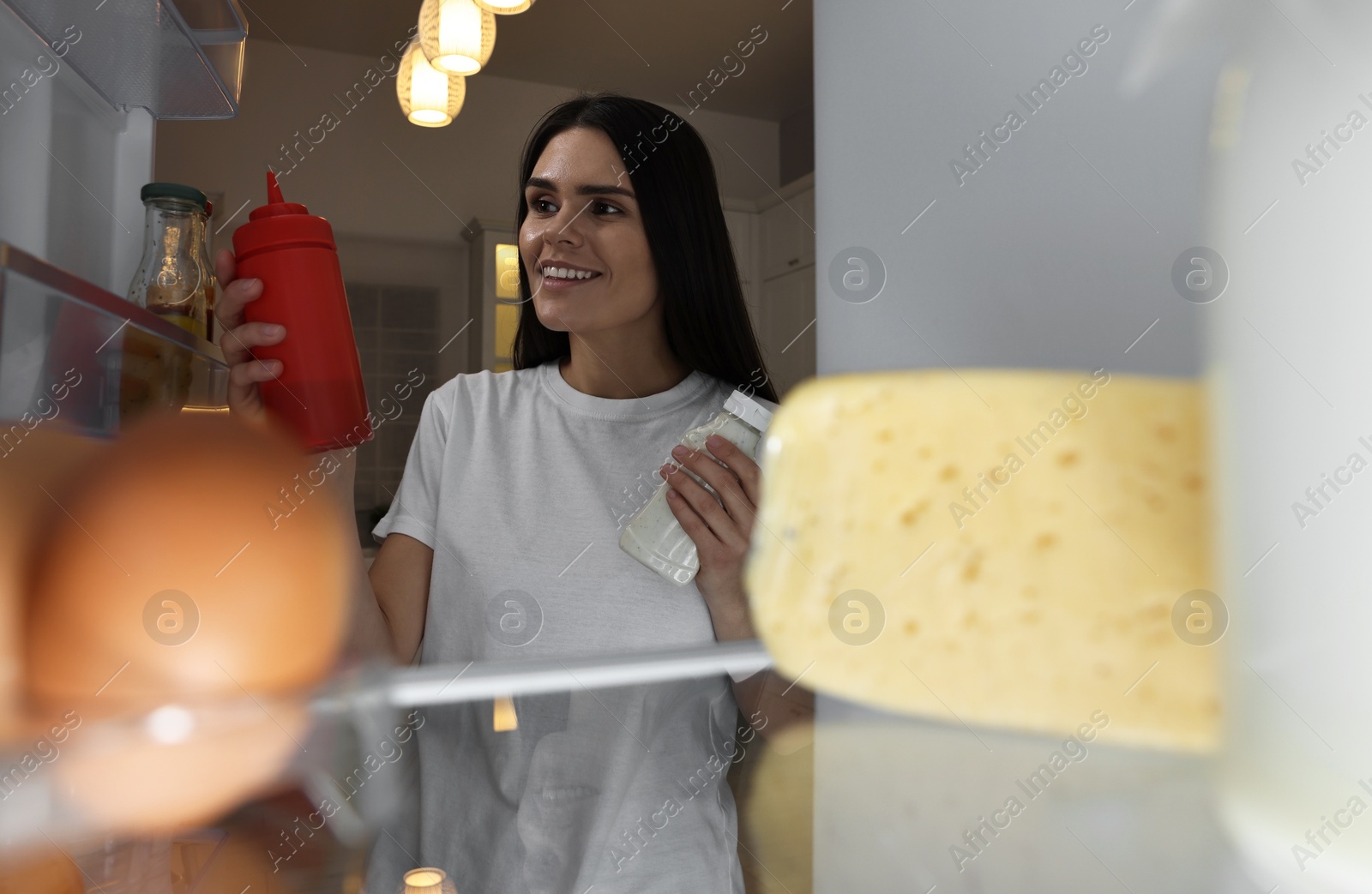 Photo of Young woman taking ketchup and mayonnaise out of refrigerator in kitchen at night, view from inside