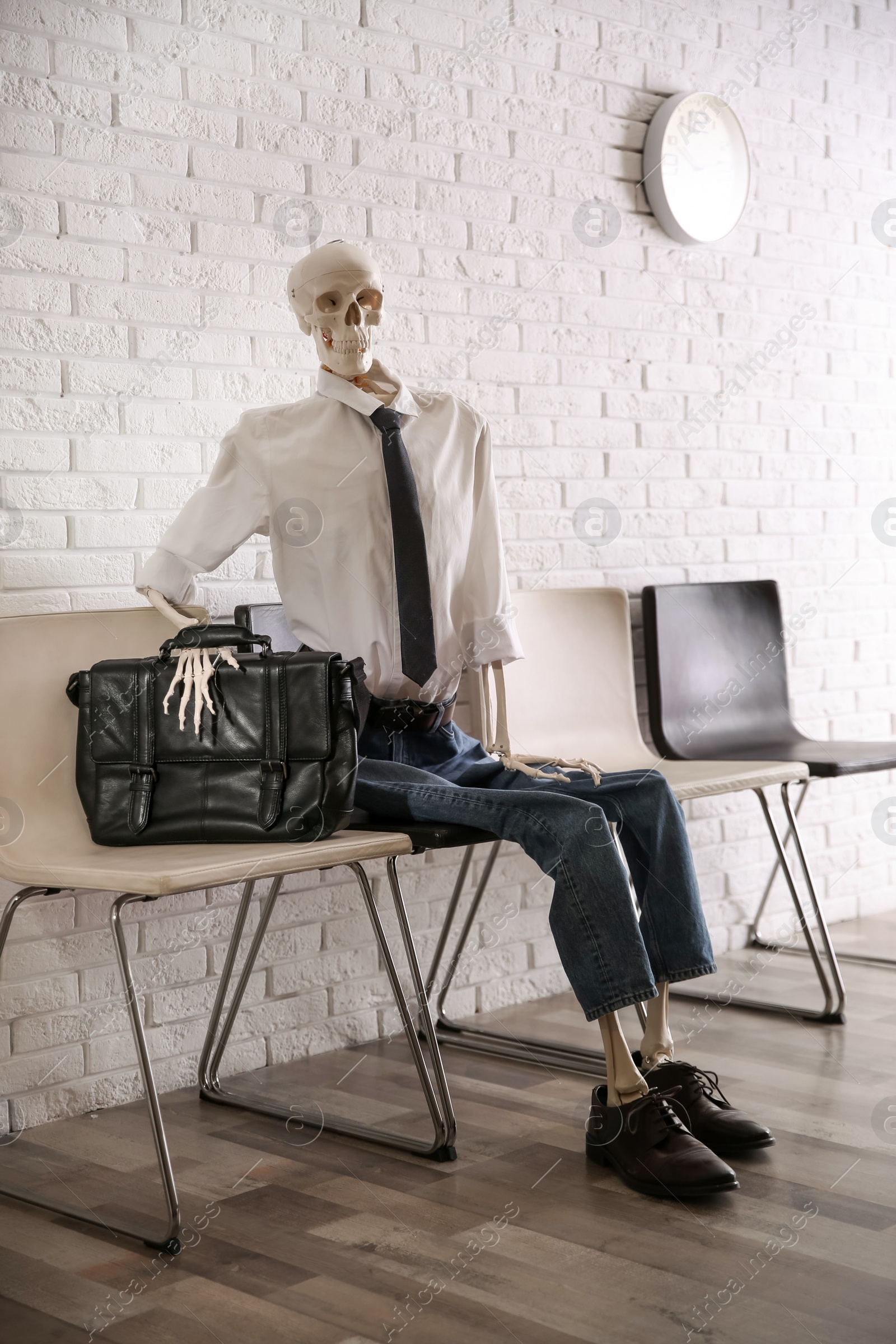 Photo of Human skeleton in office wear sitting on chair near brick wall indoors