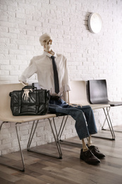 Human skeleton in office wear sitting on chair near brick wall indoors