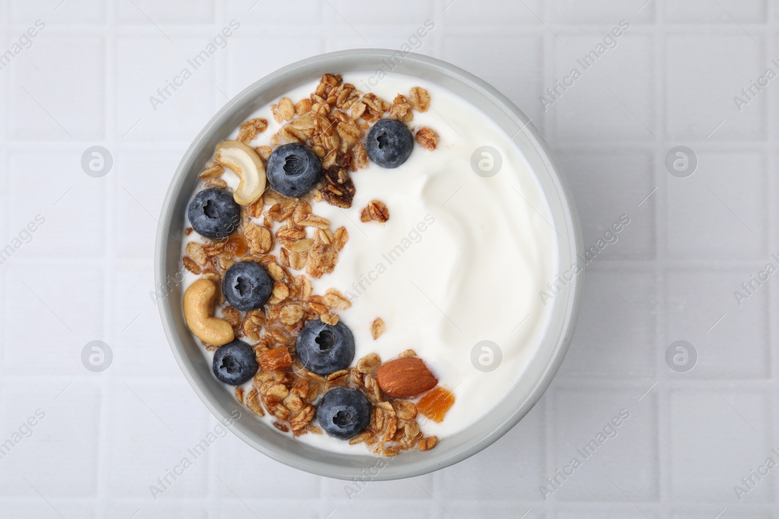 Photo of Bowl with yogurt, blueberries and granola on white tiled table, top view