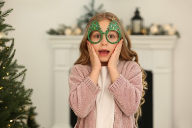 Emotional little girl wearing glasses in shape of Christmas tree at home