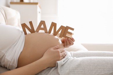 Pregnant woman with wooden letters on belly indoors, closeup. Choosing baby name