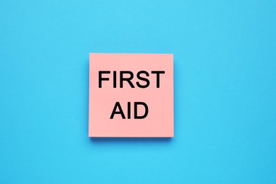 Photo of Pink paper note with words First Aid on light blue background, top view