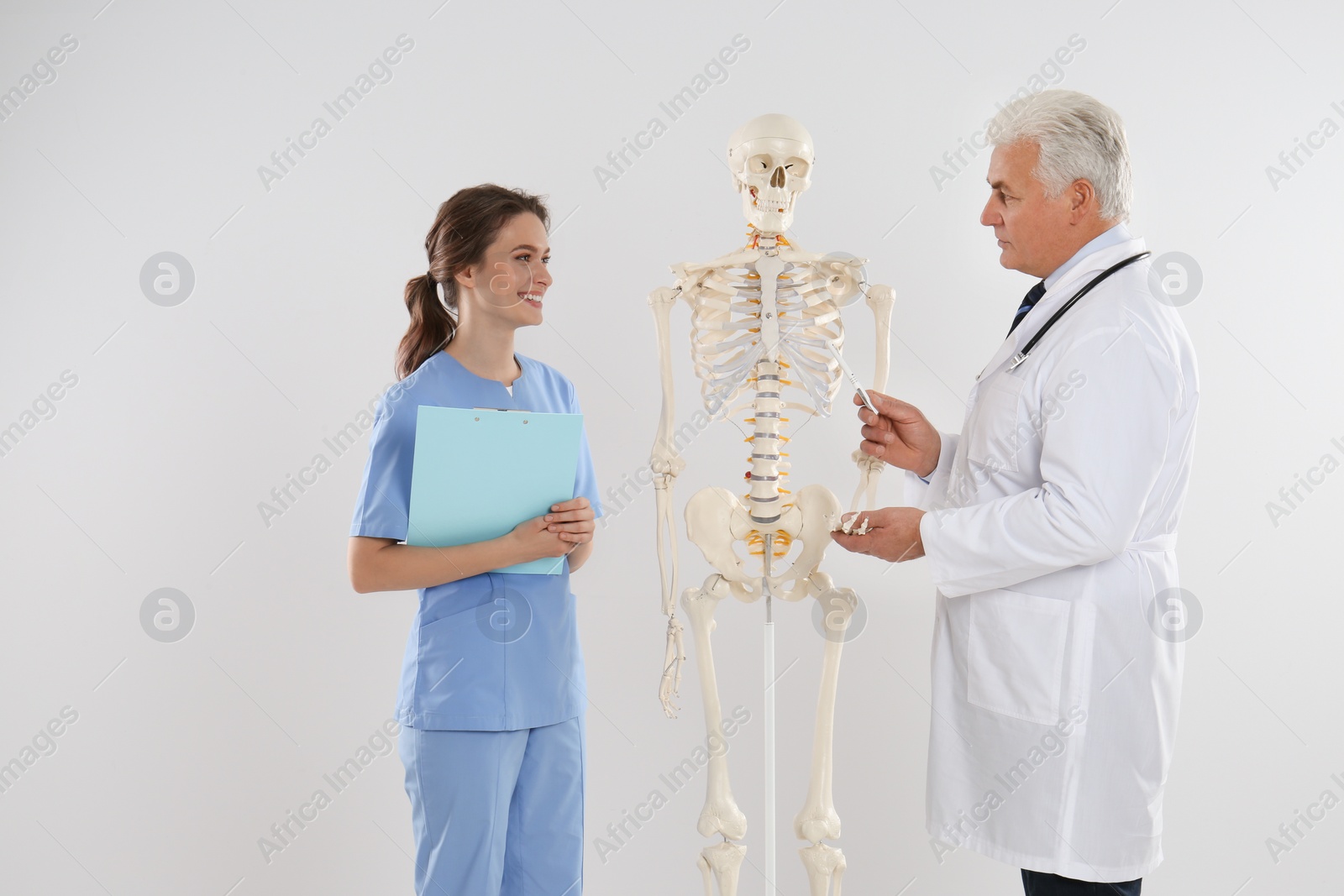 Photo of Professional orthopedist with human skeleton model teaching medical student against light background