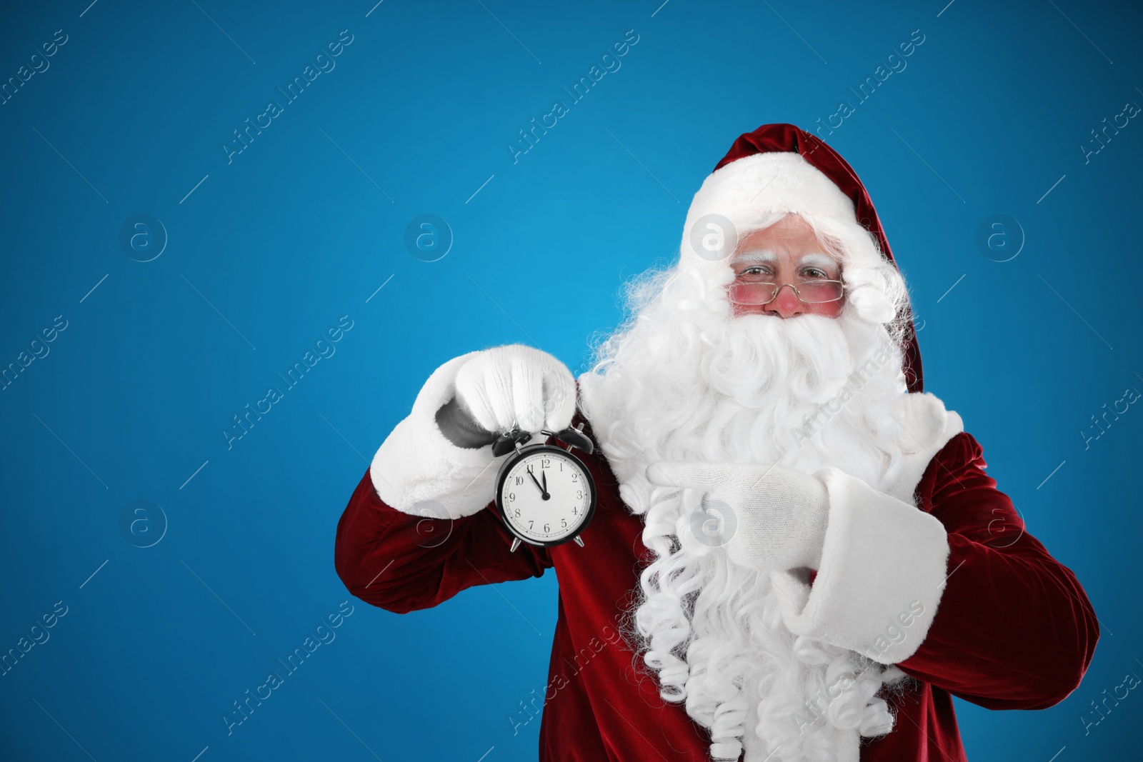 Photo of Santa Claus holding alarm clock on blue background, space for text. Christmas countdown