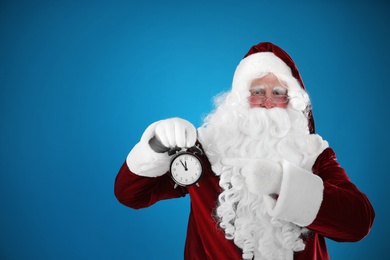 Photo of Santa Claus holding alarm clock on blue background, space for text. Christmas countdown