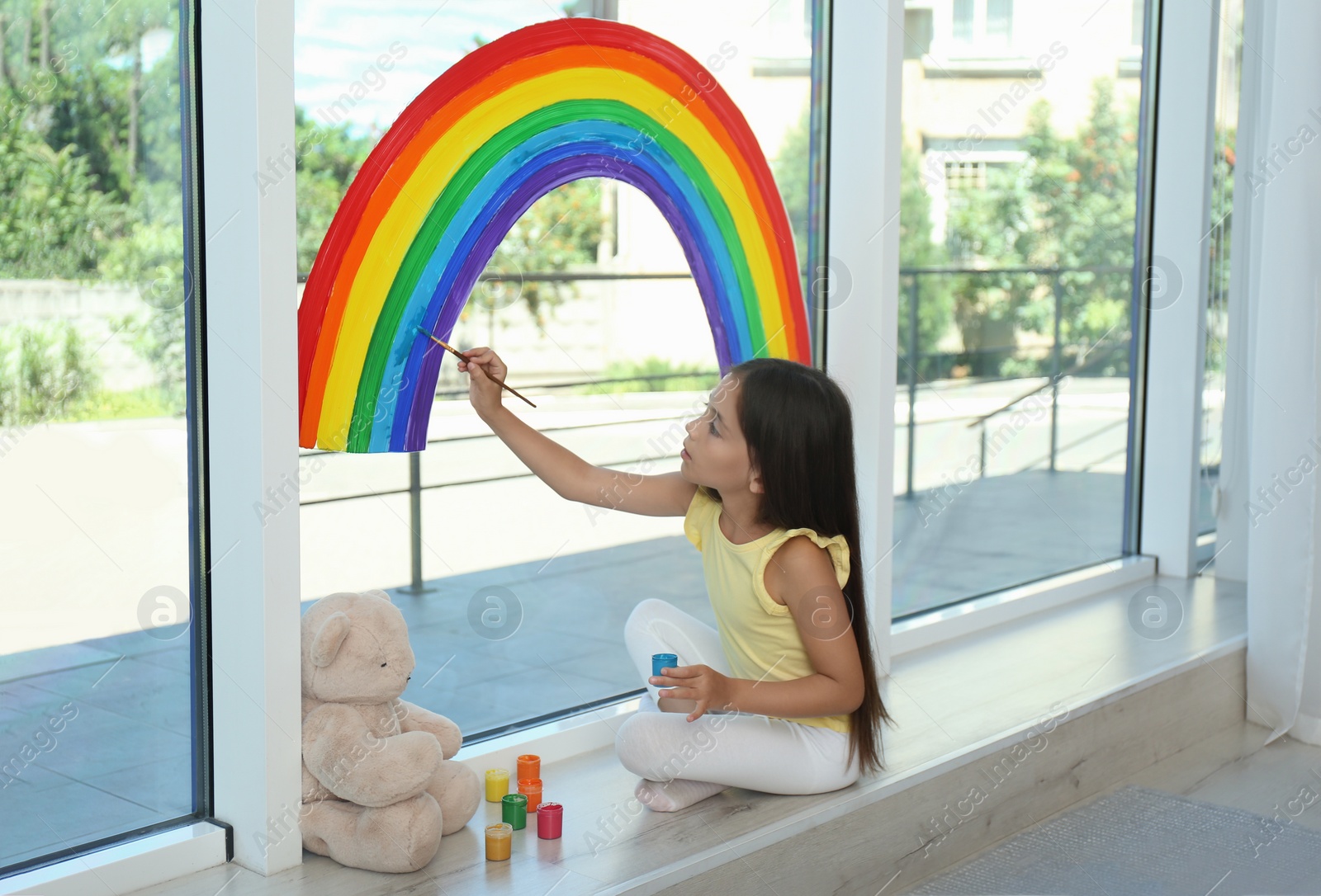 Photo of Little girl drawing rainbow on window with paints indoors. Stay at home concept