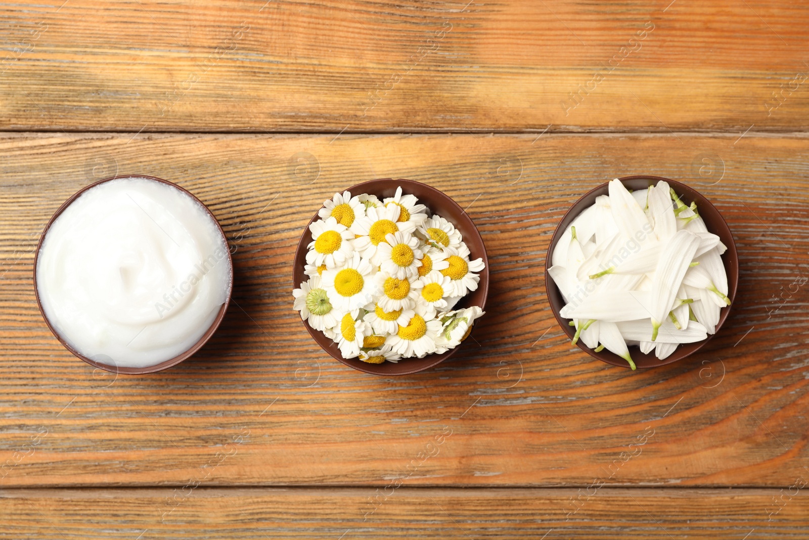 Photo of Bowls of cosmetic cream, chamomiles and petals on wooden table, flat lay