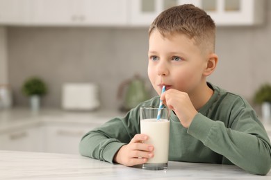 Photo of Cute boy drinking fresh milk from glass at white table in kitchen, space for text