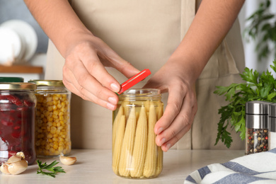 Photo of Woman closing jar with pickled corn cobs at white table, closeup