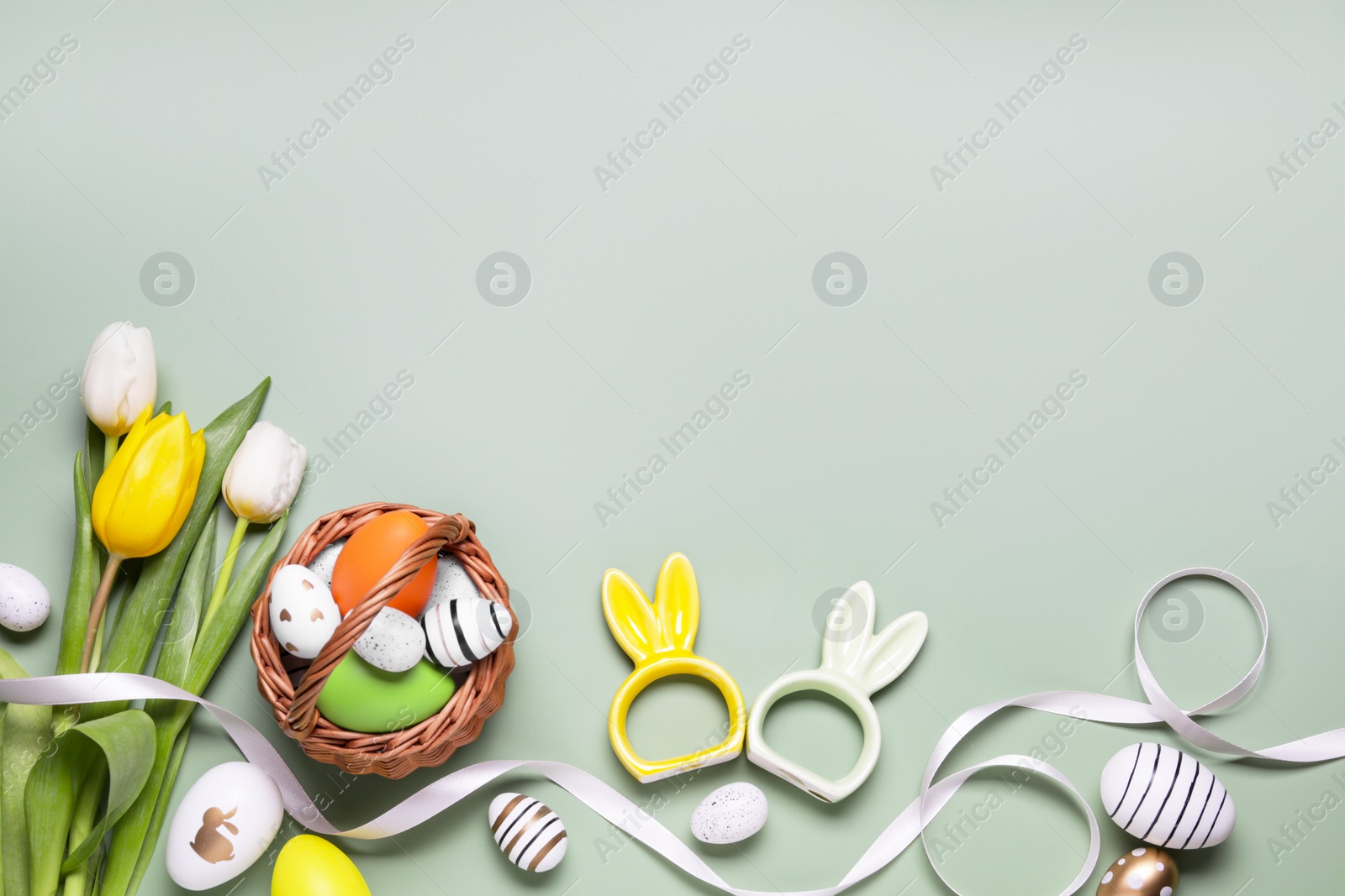 Photo of Flat lay composition with Easter eggs in basket and tulip flowers on pale olive background. Space for text