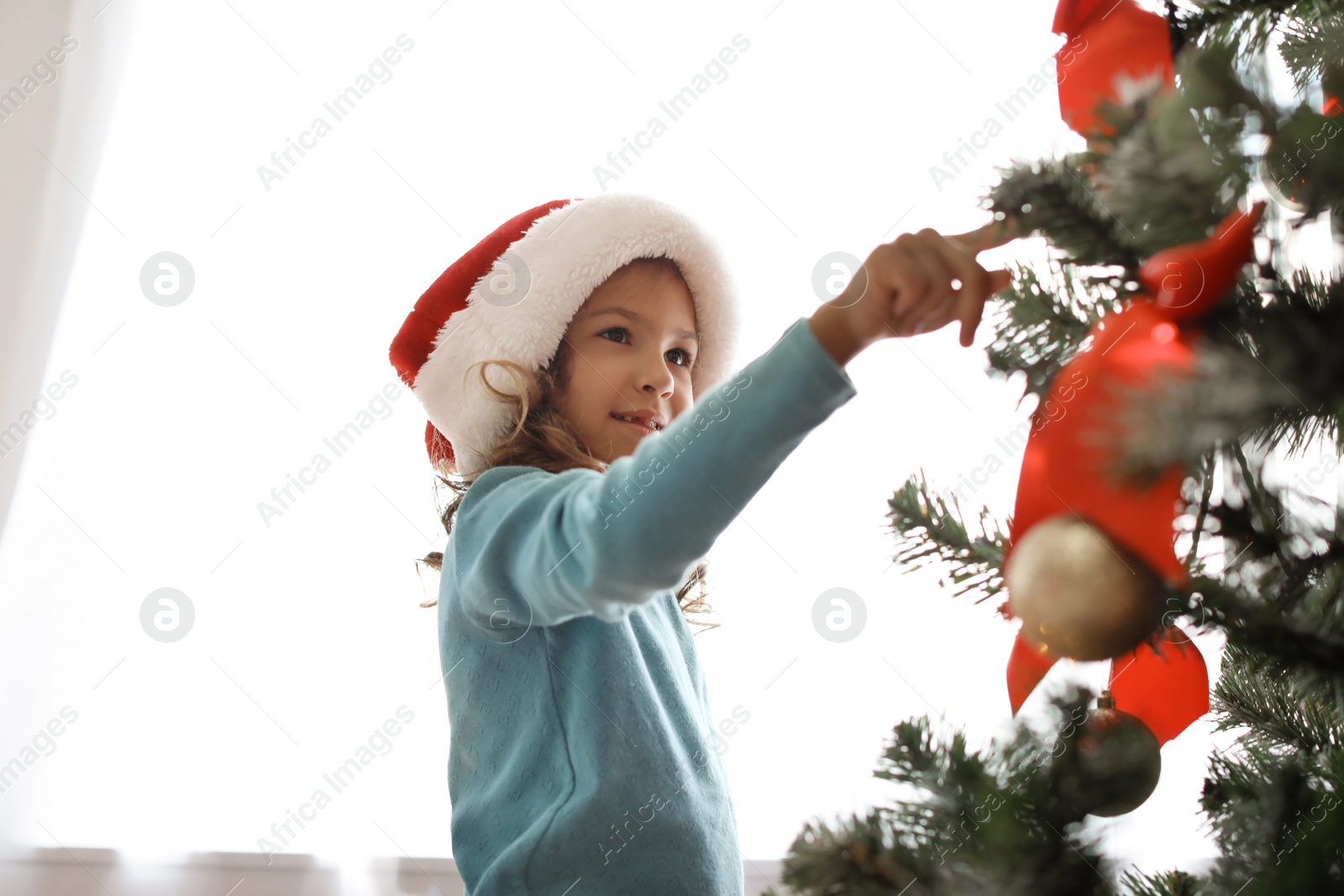 Photo of Cute little child in Santa hat near Christmas tree at home