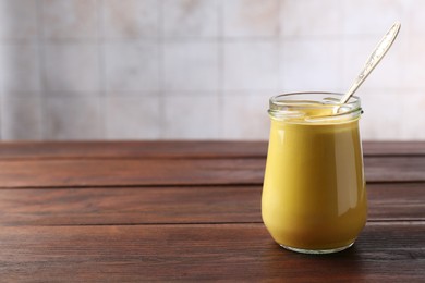 Tasty mustard sauce and spoon in glass jar on wooden table, space for text