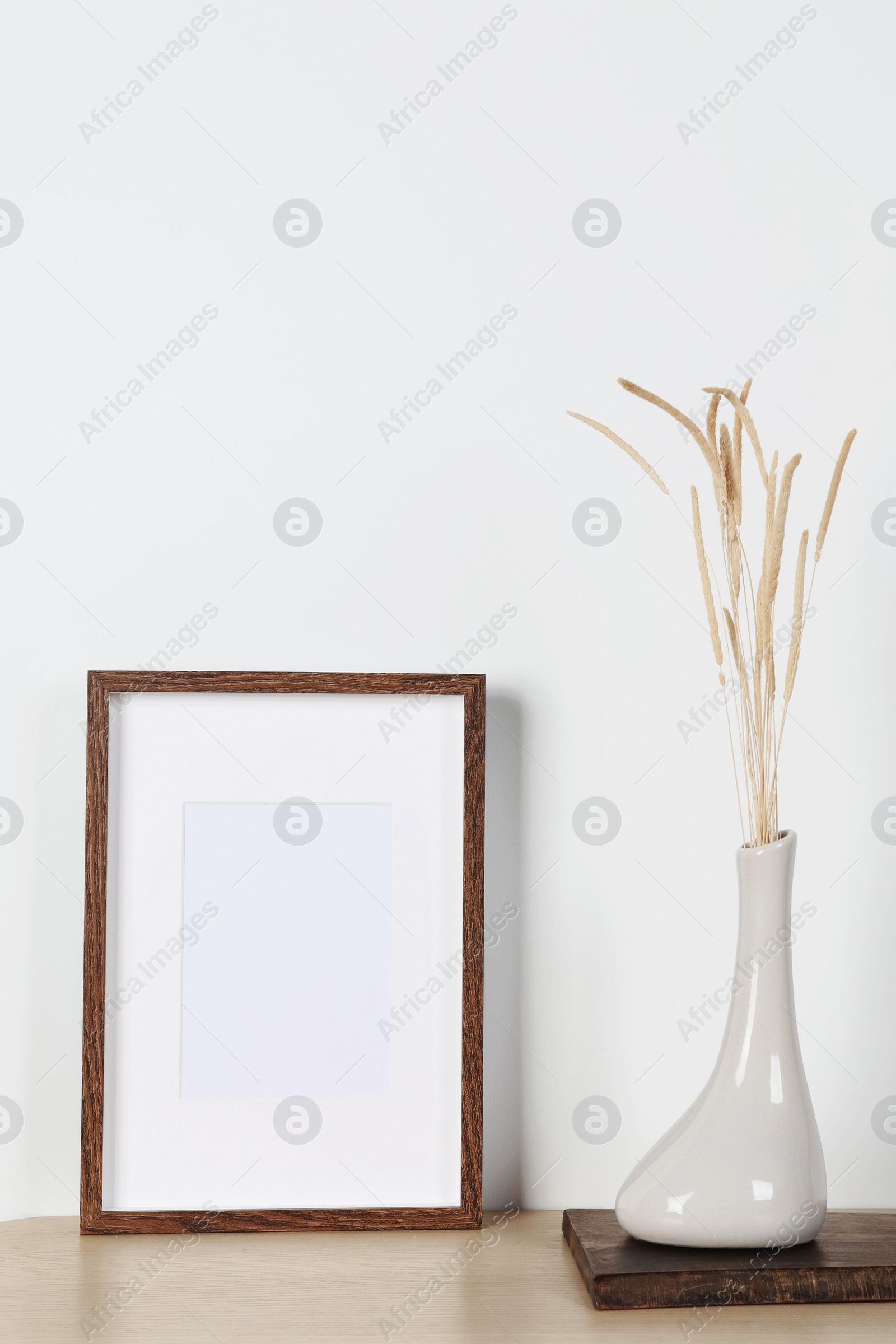 Photo of Empty photo frame and vase with dry decorative spikes on wooden table. Mockup for design