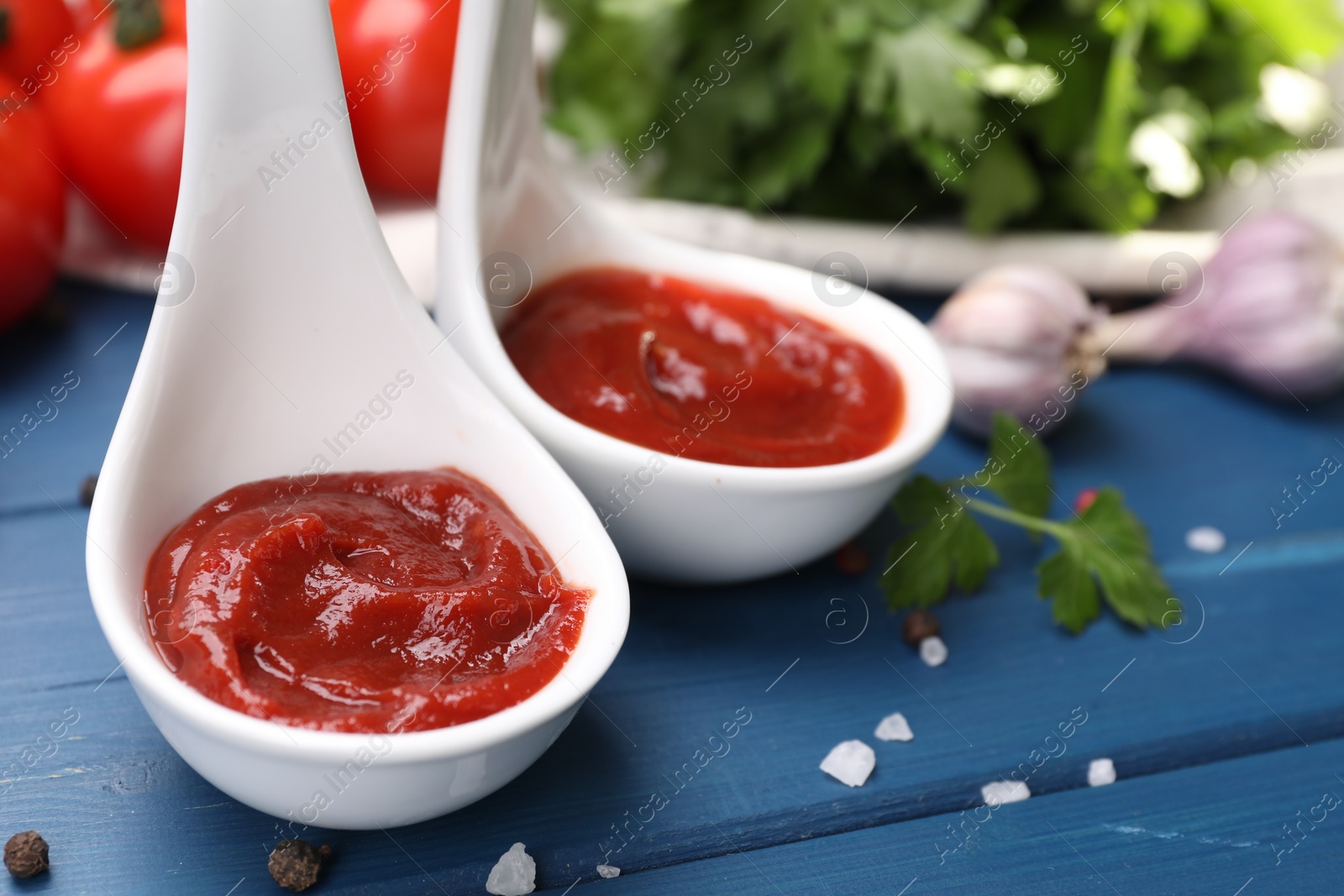 Photo of Organic ketchup in spoons and spices on blue wooden table, closeup. Tomato sauce