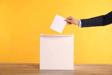 Photo of Woman putting her vote into ballot box on wooden table against orange background, closeup