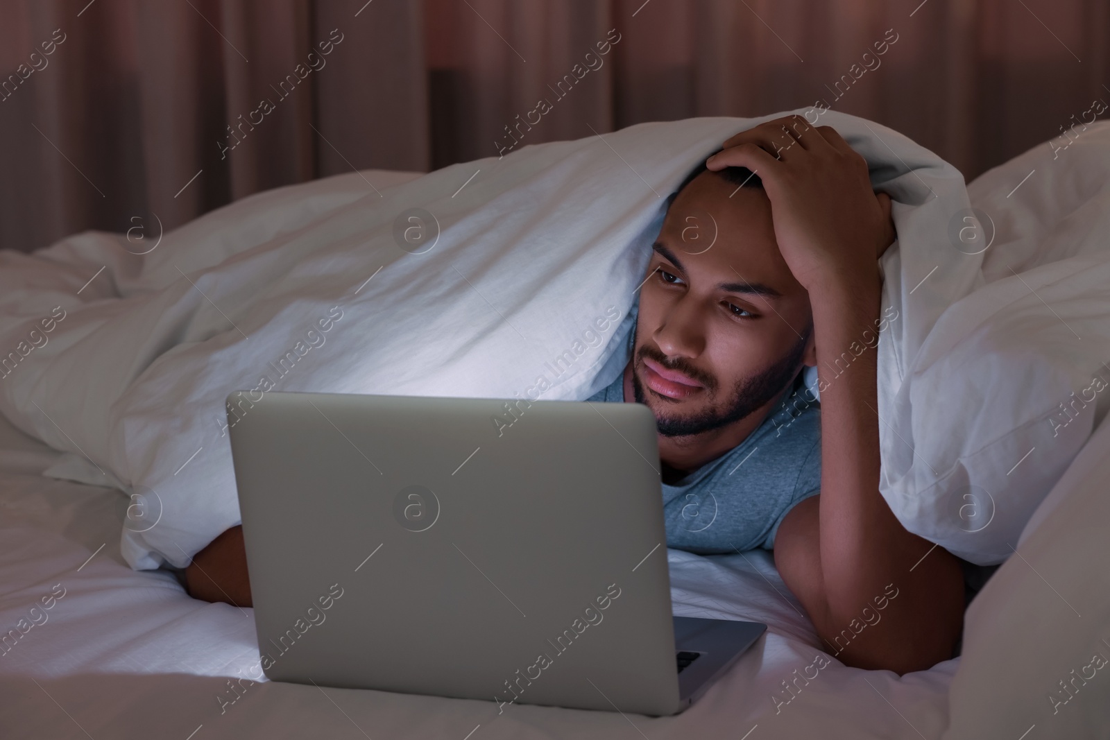 Photo of Young man using laptop under blanket in bed at night. Internet addiction