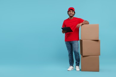 Happy young courier with clipboard and stack of parcels on light blue background, space for text