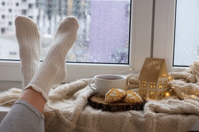 Photo of Woman in knitted socks relaxing near window at home, closeup. Space for text