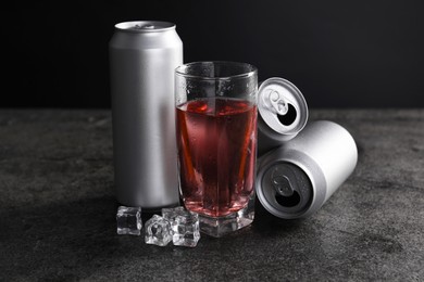 Photo of Energy drink in glass, aluminium cans and ice cubes on grey table