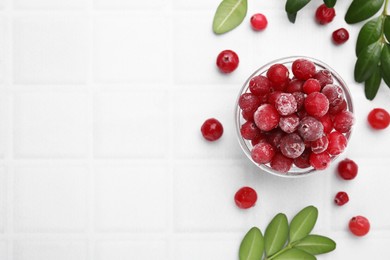 Photo of Frozen red cranberries in bowl and green leaves on white table, flat lay. Space for text