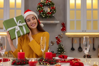 Photo of Happy young woman in Santa hat with Christmas gift at table in kitchen