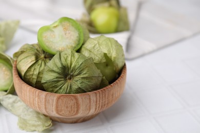 Fresh green tomatillos with husk in bowl on white tiled table, closeup. Space for text