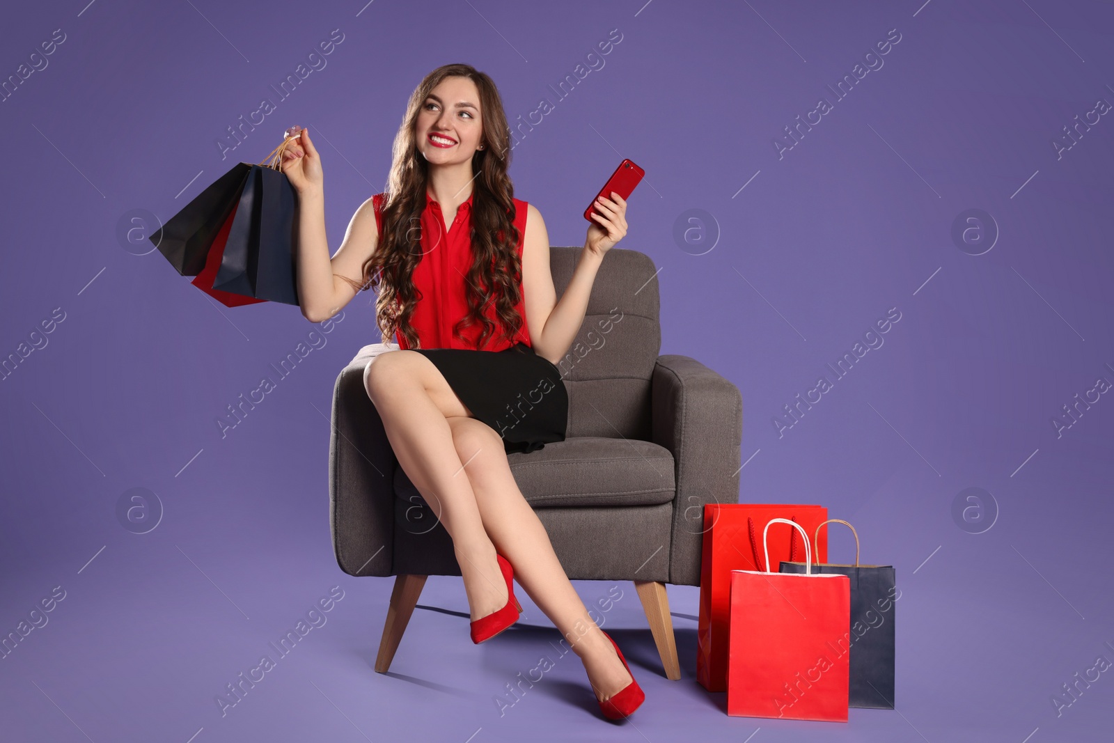 Photo of Happy woman holding paper shopping bags and smartphone on armchair against purple background