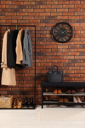 Stylish hallway with coat rack, mirror and shoe storage bench near brick wall. Interior design