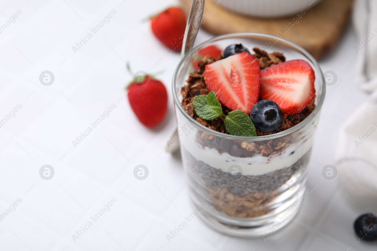 Photo of Tasty granola with berries, yogurt and chia seeds in glass on white table, closeup. Space for text