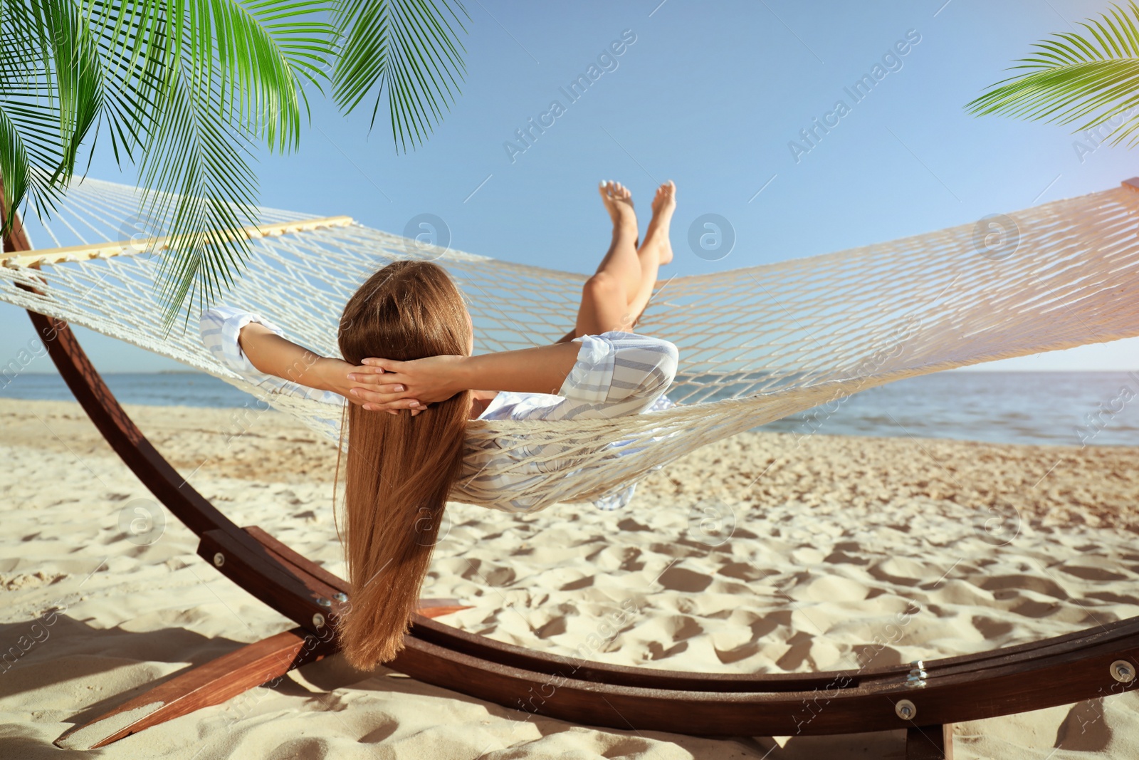 Image of Woman relaxing in hammock under green palm leaves on sunlit beach