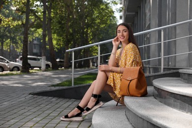 Photo of Beautiful young woman with stylish backpack sitting on stairs outdoors