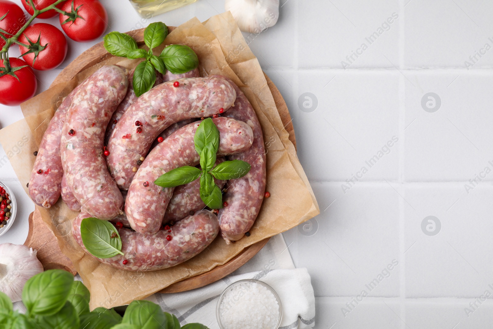 Photo of Raw homemade sausages, basil leaves, tomatoes and peppercorns on white tiled table, flat lay. Space for text