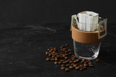 Glass cup with drip coffee bag and beans on black slate table, closeup. Space for text