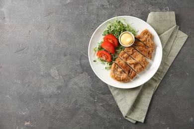 Tasty cut schnitzel served with sauce, microgreens and tomato on grey textured table, top view. Space for text