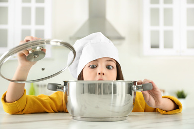 Photo of Surprised little girl wearing chef hat with pot in kitchen