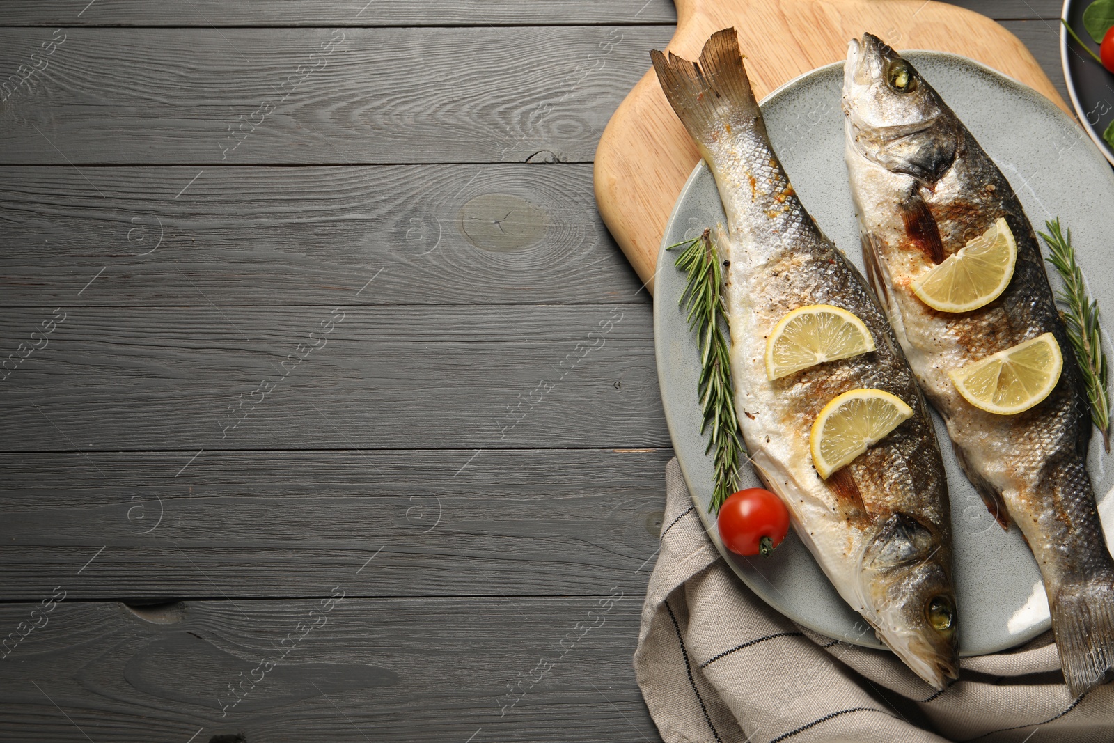 Photo of Delicious baked fish, tomato, lemon and rosemary on grey wooden table, top view. Space for text