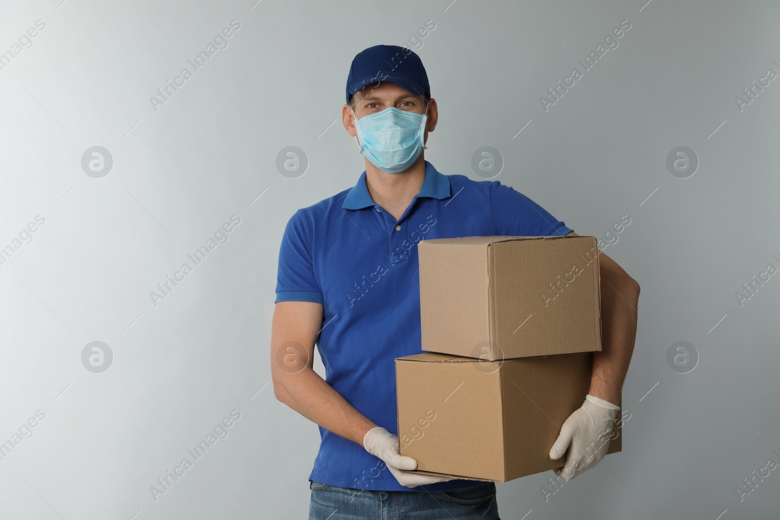 Photo of Courier in protective mask and gloves holding cardboard boxes on light background. Delivery service during coronavirus quarantine