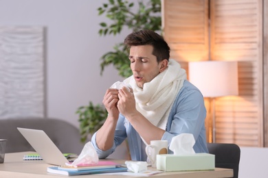 Sad exhausted man with tissue suffering from cold while working with laptop at table