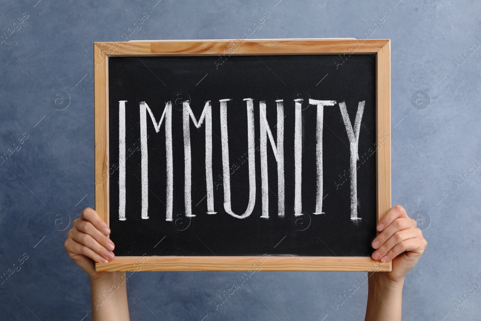 Photo of Woman holding small chalkboard with word Immunity on blue background, closeup