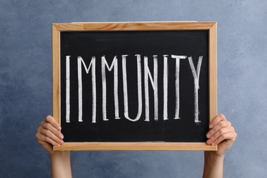 Photo of Woman holding small chalkboard with word Immunity on blue background, closeup