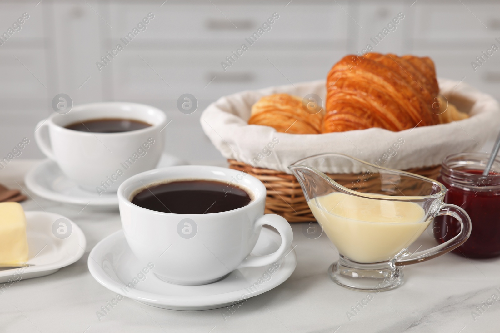 Photo of Breakfast time. Fresh croissants, coffee, sweetened condensed milk and jam on white table, closeup