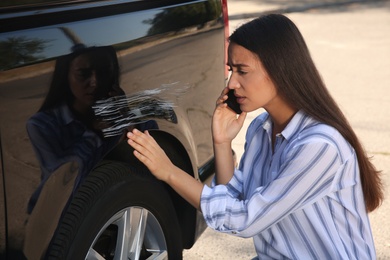 Photo of Stressed woman talking on phone near car with scratch outdoors