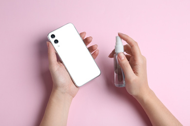 Photo of Woman spraying antiseptic onto smartphone on pink background, top view