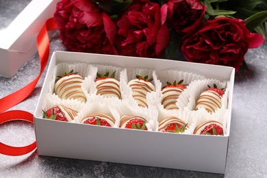 Photo of Delicious chocolate covered strawberries in box and flowers on light grey table